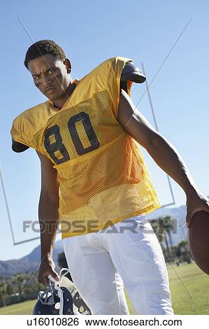 Stock Photograph - Football player holding helmet and ball standing on field, low angle view, portrait, (portrait). Fotosearch