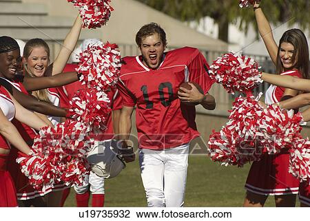 Stock Image - Football player running between cheerleaders on field. Fotosearch