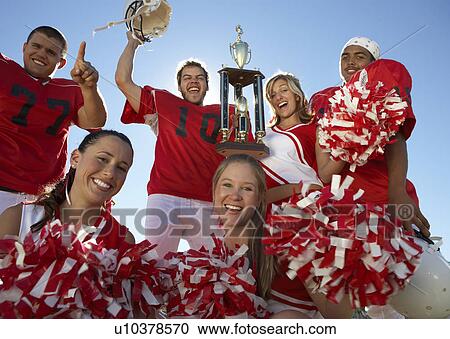 Stock Image - Football players and cheerleaders hoisting trophy, low angle view, portrait, (portrait). Fotosearch
