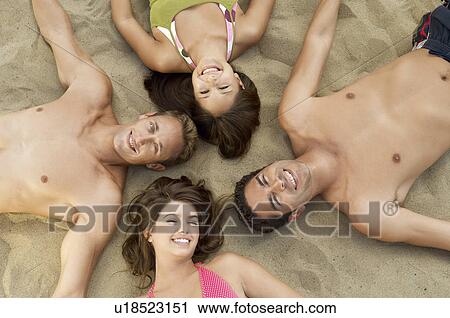 Stock Image - Four young adults lying down in circle on sand, smiling, portrait, view from above. Fotosearch