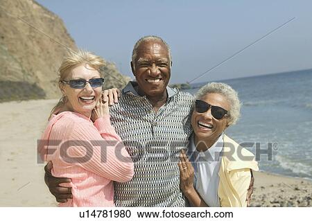 Stock Image - Friends embracing on beach, smiling, (portrait). Fotosearch
