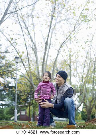 Stock Image - Girl (3-4) standing with father, squatting, in park. Fotosearch