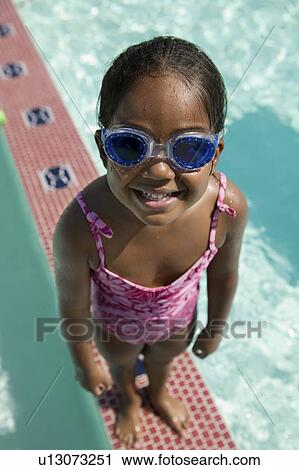 Stock Image - Girl (5-6) Wearing swim Goggles standing on Edge of Swimming Pool, elevated view portrait.. Fotosearch