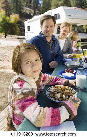 Stock Image - Girl eating at picnic table with family in campground. Fotosearch