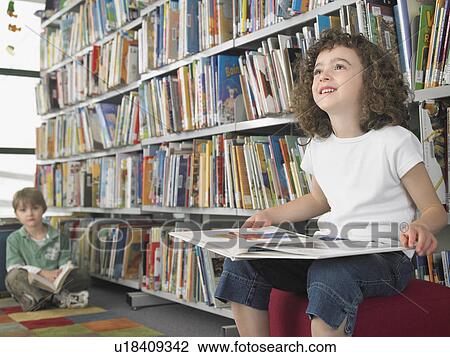 Stock Image - Girl holding open book, sitting in library. Fotosearch