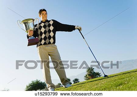 Stock Photo - Golfer holding trophy standing on golf course, (portrait). Fotosearch