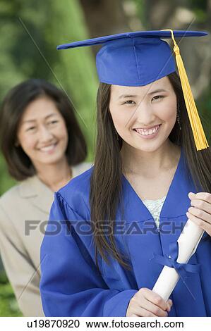 Stock Image - Graduate holding diploma with mother behind, outside, portrait. Fotosearch