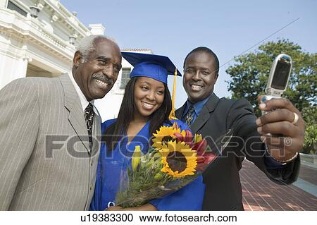 Stock Image - Graduate with father and grandfather taking picture with cell phone outside university. Fotosearch