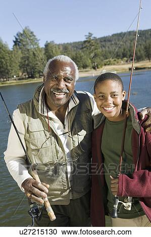 Stock Image - Grandfather and grandson holding fishing rods by lake, smiling, (portrait). Fotosearch