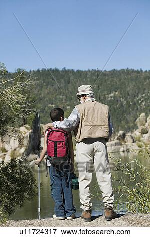Stock Photo - Grandfather and grandson standing by lake, holding fishing rods, back view. Fotosearch