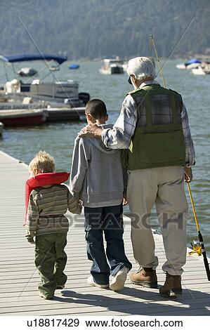 Stock Photo - Grandfather and two grandsons holding fishing rods, walking on pier, back view. Fotosearch