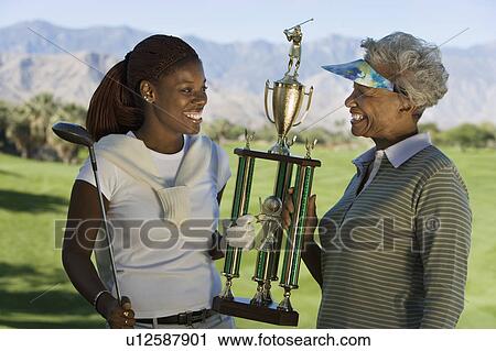 Stock Image - Grandmother and granddaughter holding golf trophy, smiling. Fotosearch