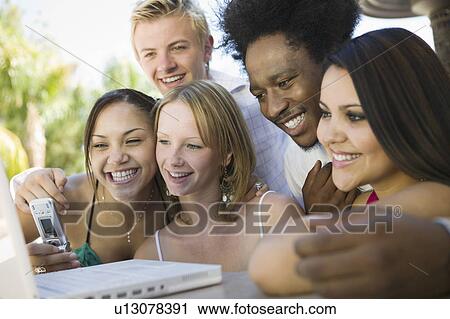 Stock Image - Group of friends at back yard table using laptop and cell phone, close up. Fotosearch