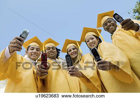 Stock Image - Group of graduates taking pictures with cell phone outside, low angle view. Fotosearch