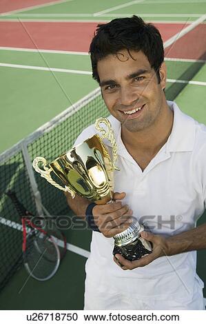 Stock Image - Male Tennis Player standing on tennis court holding trophy, portrait, high angle view. Fotosearch