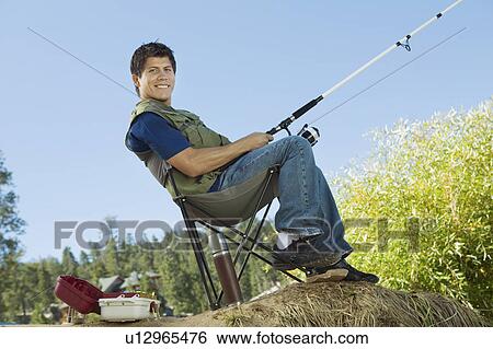Stock Photograph - Man fly fishing, sitting on collapsible chair, (portrait). Fotosearch