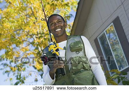 Stock Image - Man holding fishing rod in front of house, smiling, (low angle view). Fotosearch