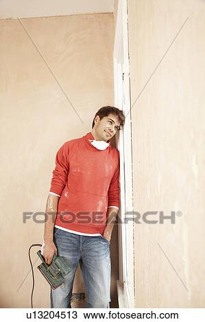Stock Image - Man holding sanding tool, leaning against wall of unrenovated room. Fotosearch