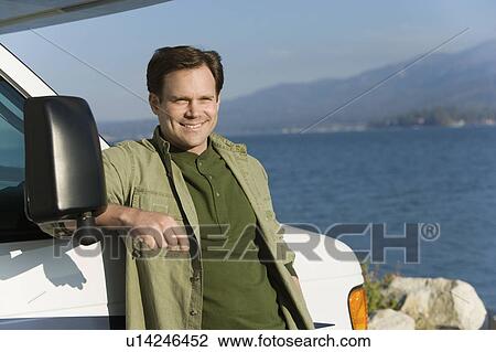 Stock Image - Man leaning on RV at lake. Fotosearch