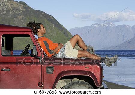 Stock Image - Man lying on hood of jeep near mountain lake. Fotosearch
