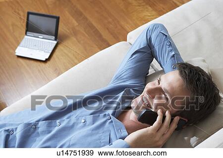 Stock Photo - Man lying on sofa using mobile phone, laptop in background, view from above.. Fotosearch