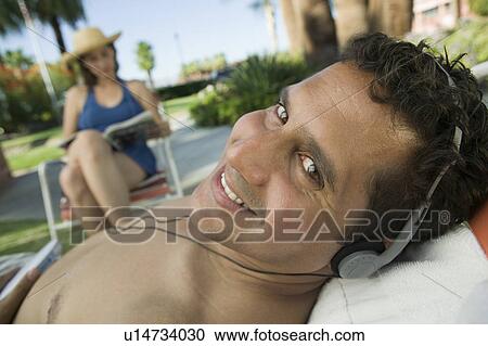 Stock Image - Man Lying on sunlounger Listening to headphones, portrait.. Fotosearch