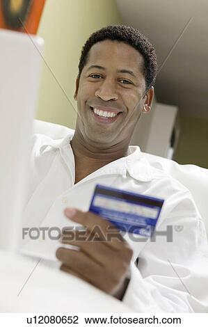 Stock Image - Man on sofa Using Credit Card to Make Purchase with Laptop, portrait, low angle view. Fotosearch