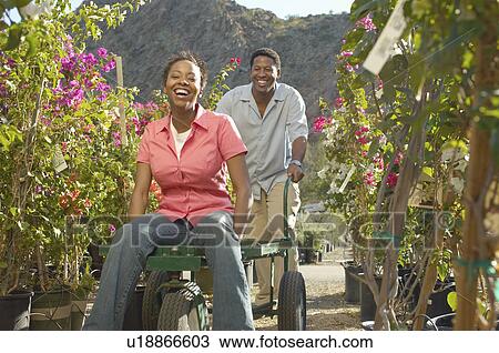 Stock Image - Man pushing woman on cart at plant nursery. Fotosearch