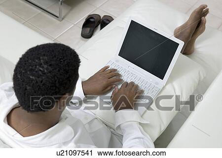 Stock Image - Man reclining on sofa using laptop, high angle view. Fotosearch