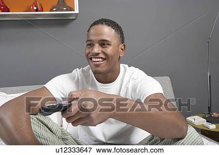 Stock Photo - Man sitting on bed using remote control. Fotosearch