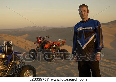 Stock Photo - Man standing by quad bikes in desert at sunset. Fotosearch