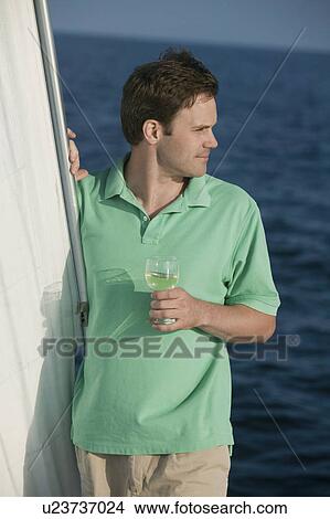 Picture - Man standing on sailboat, holding glass of white wine. Fotosearch