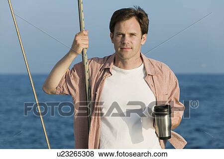 Stock Photo - Man standing on sailboat, holding coffee mug, (portrait). Fotosearch