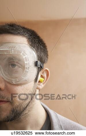 Stock Photography - Man wearing protective eye goggles and earplugs indoor, close up. Fotosearch