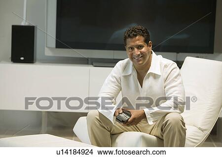 Man with remote control sitting in front of Large Screen TV in living room, portrait View Large Photo Image Picture - Man with remote control sitting in front of Large Screen TV in living room, portrait. Fotosearch