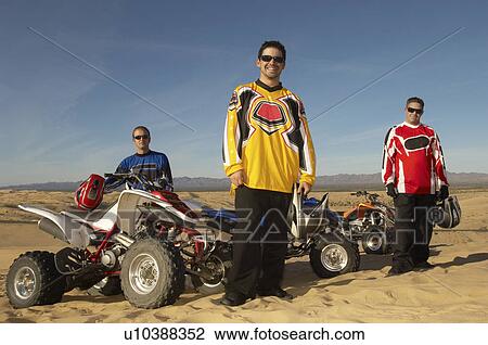 Stock Image - Men standing by quad bikes in desert. Fotosearch
