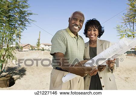 Stock Photograph - Middle-aged couple holding blueprints in vacant lot, portrait. Fotosearch