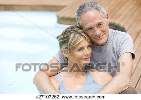 Stock Image - Middle-aged couple sitting embracing by swimming pool, portrait. Fotosearch