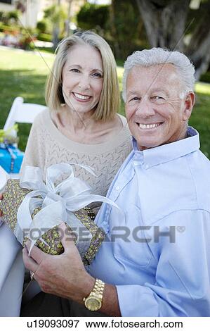 Stock Photo - Middle-aged Couple sitting, Holding a Gift outside. Fotosearch