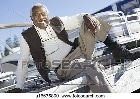Stock Image - Middle-aged fisherman sitting on pier, (portrait). Fotosearch