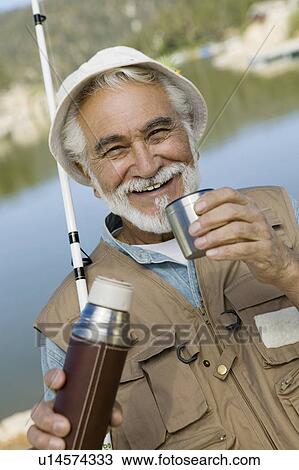 Stock Image - Middle-aged man fishing, holding vacuum flask, smiling, (portrait). Fotosearch