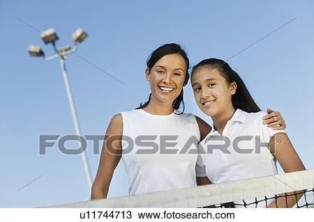 Stock Image - Mother and daughter standing at net on tennis court, portrait, low angle view. Fotosearch