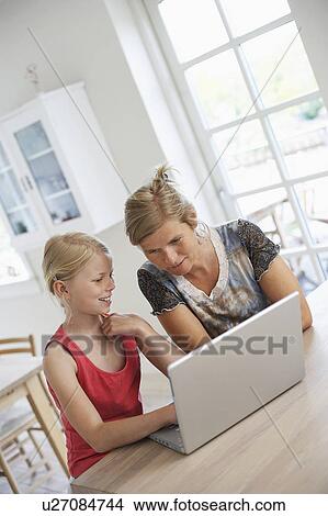 Picture - Mother using laptop with daughter at kitchen table. Fotosearch