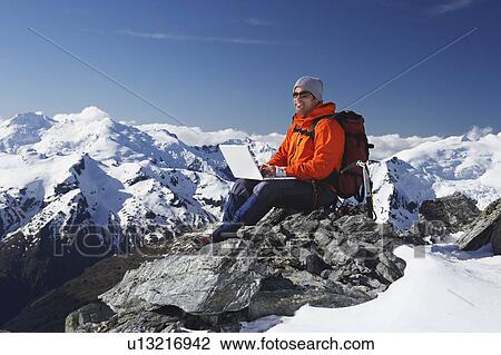 Stock Image - Mountain climber using laptop on mountain peak. Fotosearch