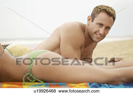 Stock Photo - Portrait of mid-adult man lying down on beach, mid section of woman on foreground. Fotosearch