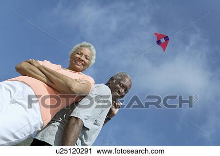 Stock Image - Senior couple flying kite, outdoors, (low angle view), (portrait). Fotosearch