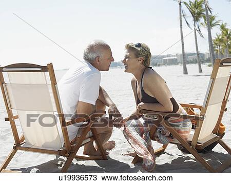 Stock Image - Senior couple on sunloungers on tropical beach, side view. Fotosearch