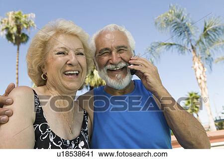 Stock Image - Senior Couple sitting outdoors, man using mobile phone, portrait.. Fotosearch