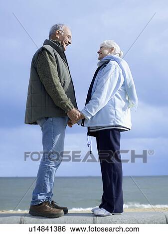 Stock Photograph - Senior couple standing on wall by water, holding hands. Fotosearch