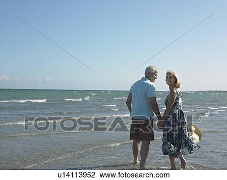 Stock Image - Senior couple walking on tropical beach, back view. Fotosearch